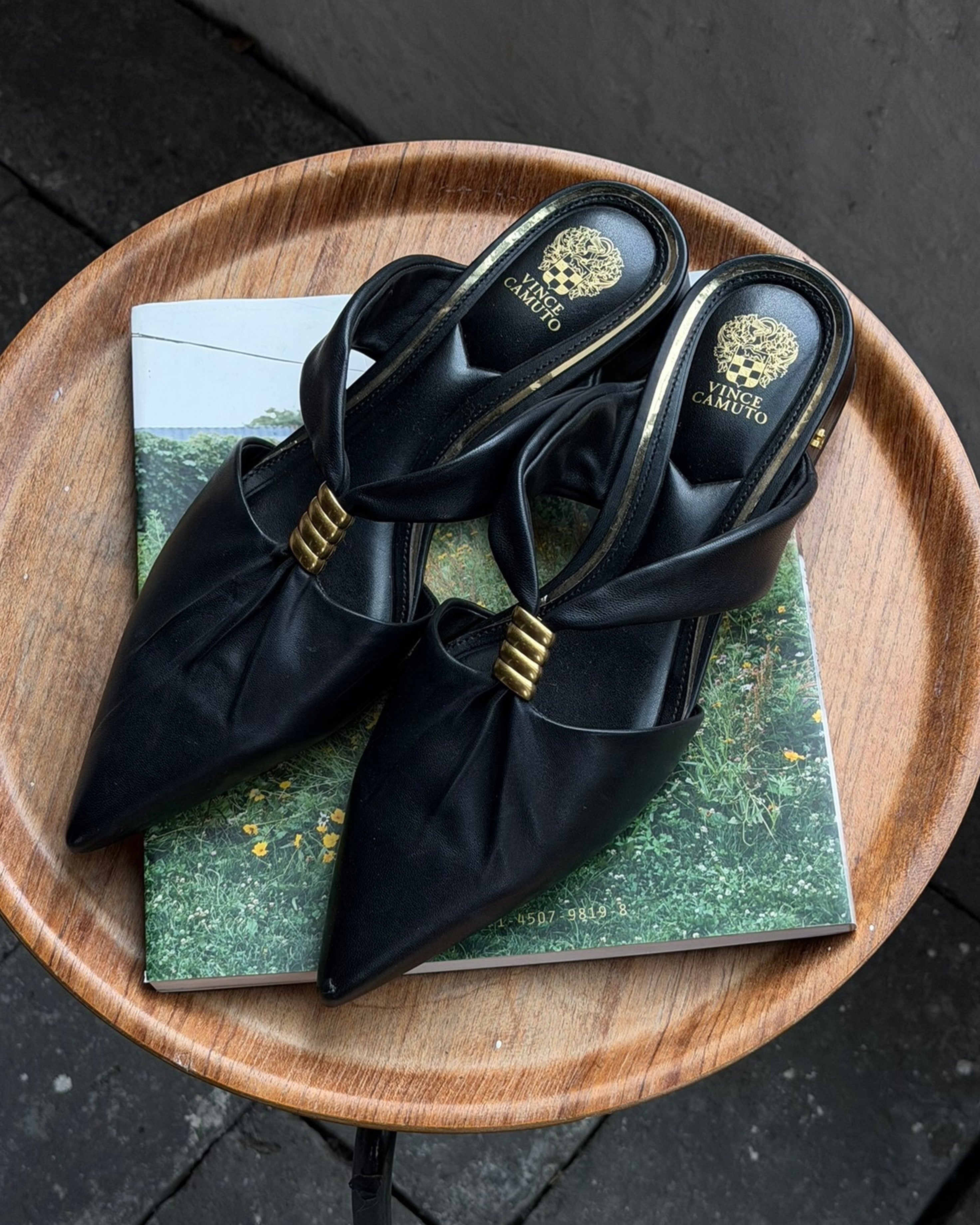 A pair of elegant black Vince Camuto Vanika Mule pointed-toe heels with gold accents rest on a wooden round table, placed atop a closed green book featuring a nature scene.