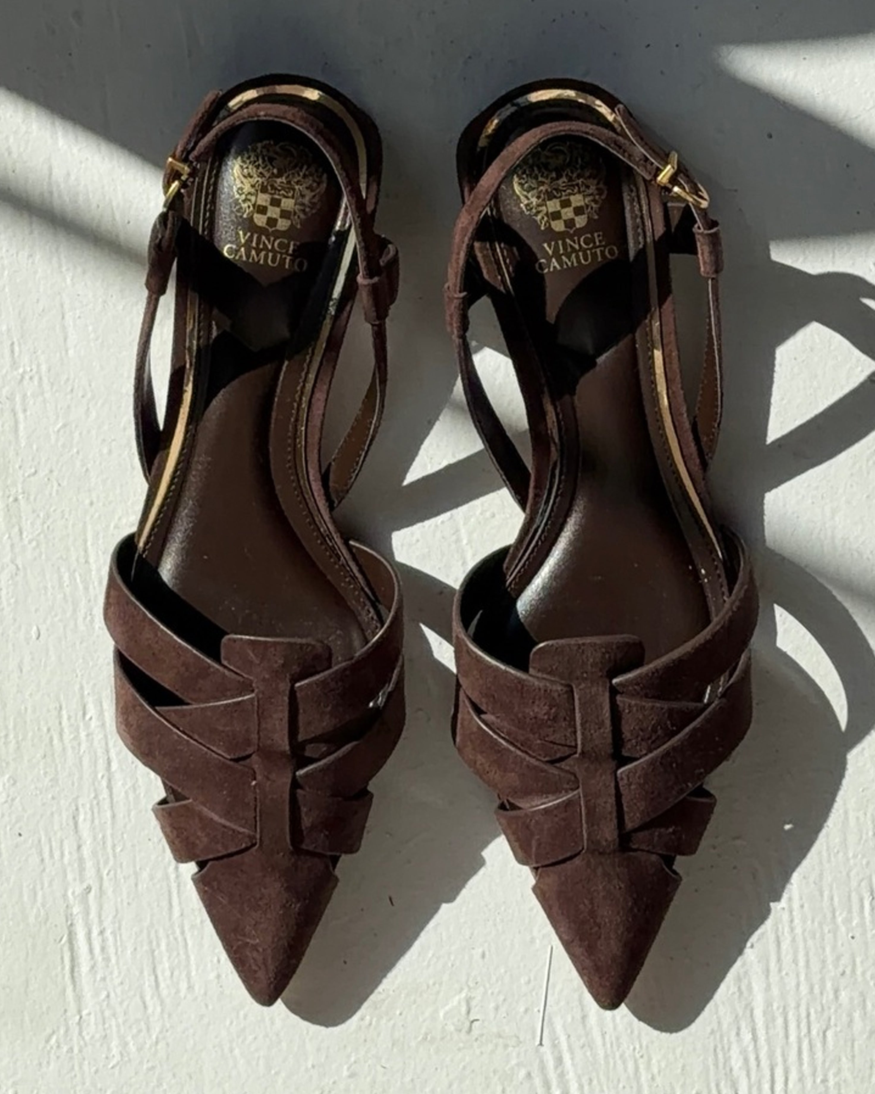 A pair of Vince Camuto Vinesha Sandals in dark brown suede with pointed toes, crisscross straps, and ankle buckles, photographed on a light surface with sunlight and shadows.