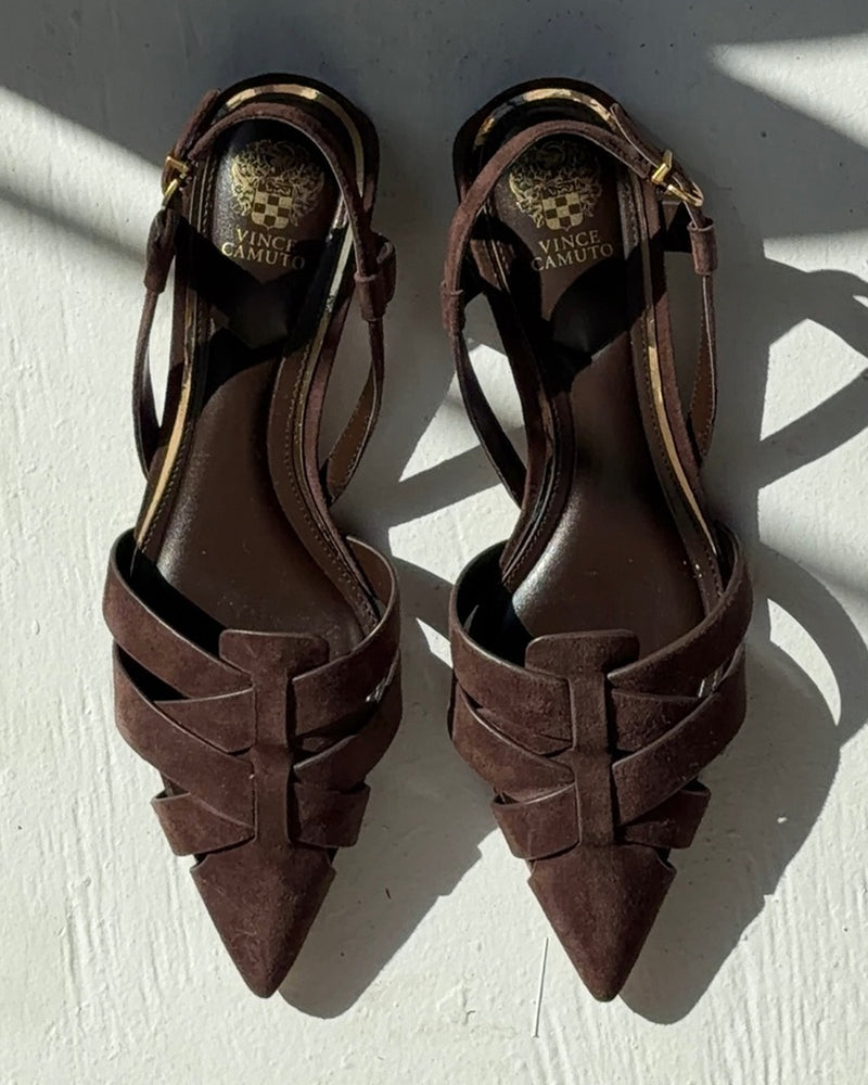 A pair of Vince Camuto Vinesha Sandals in dark brown suede with pointed toes, crisscross straps, and ankle buckles, photographed on a light surface with sunlight and shadows.