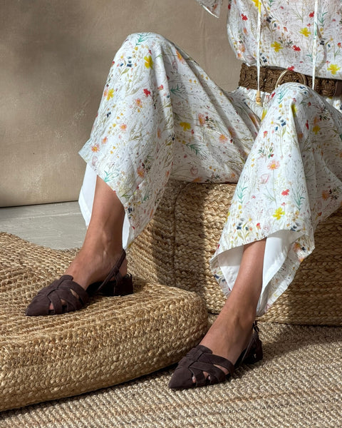 A person sits on a woven ottoman in white floral wide-leg pants and wears Vince Camuto Vinesha Sandals. The background is neutral and textured, with the upper body partially out of frame.