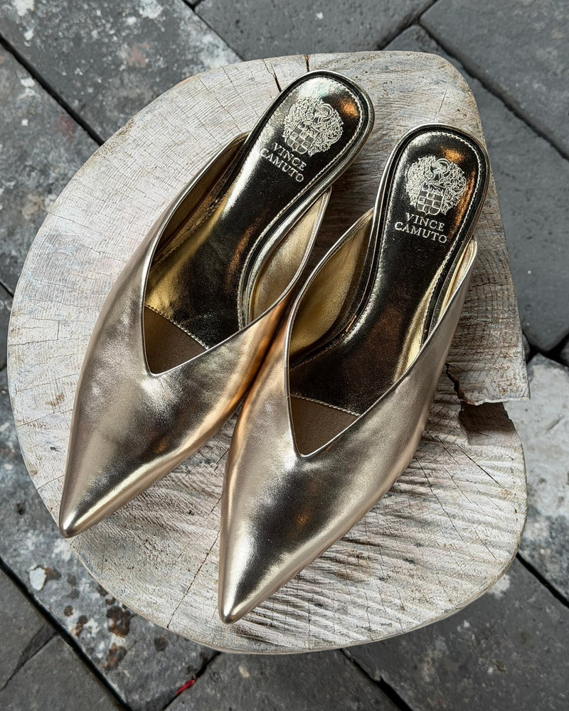 A pair of Vinia Mule gold pointed-toe slip-ons by Vince Camuto displayed on a round wooden surface over grey stone tiles.