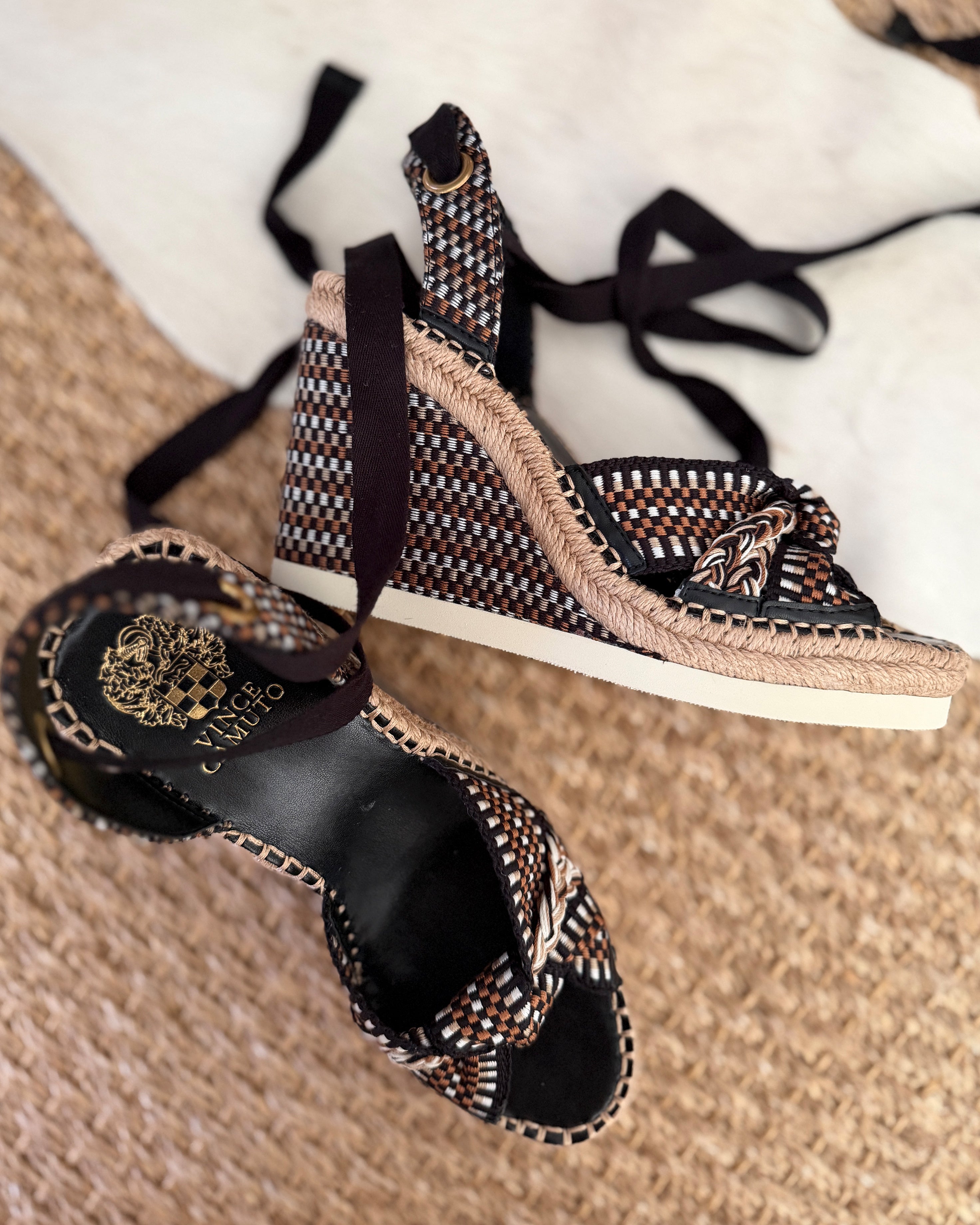 The Vince Camuto Bealie Wedge Sandal features black, brown, and beige woven straps, an ankle tie closure, and espadrille-style wedge heels, displayed on a textured beige rug.