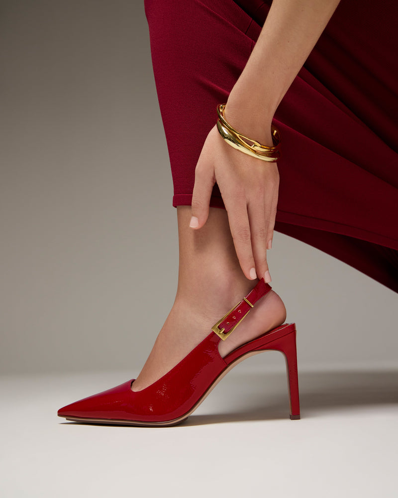 Wearing a red dress, a person adjusts the strap of the shiny red Vince Camuto Brendie Pump. A gold bracelet adorns their wrist against a plain, neutral background.