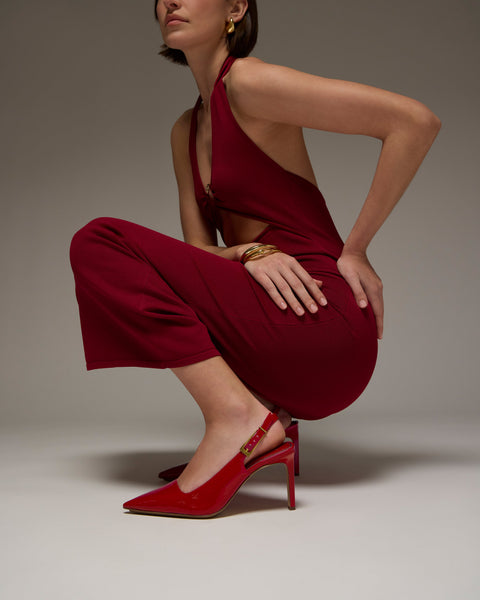 A woman poses stylishly against a neutral background, squatting in a sleeveless deep red jumpsuit and Vince Camuto Brendie Pumps. She accessorizes with gold hoop earrings and a bangle, her face partially out of the frame.