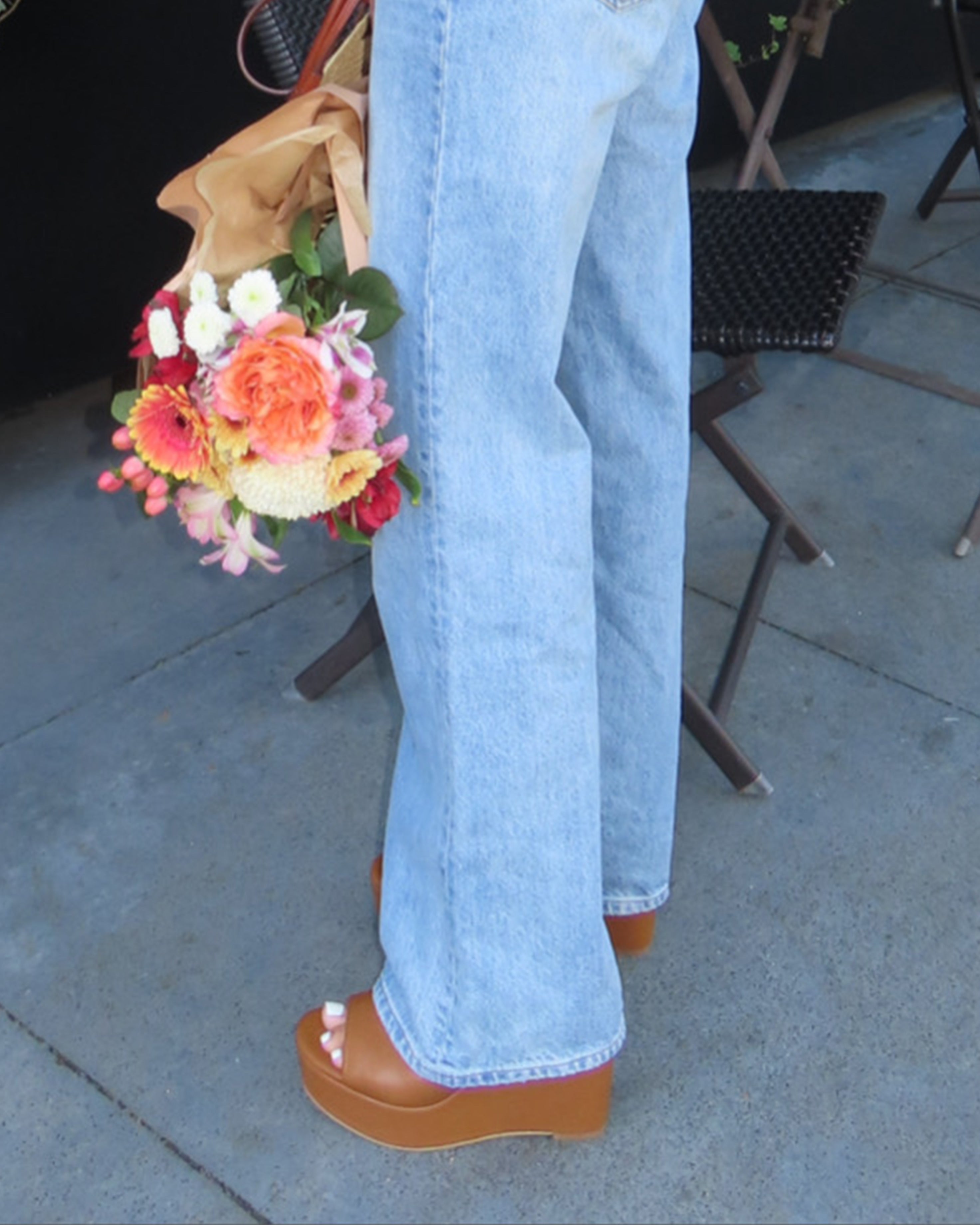 A person in light blue jeans and Vince Camuto’s Etta Wedge Sandals stands outside, holding a colorful bouquet. In the background, there’s a brown folding chair and table.