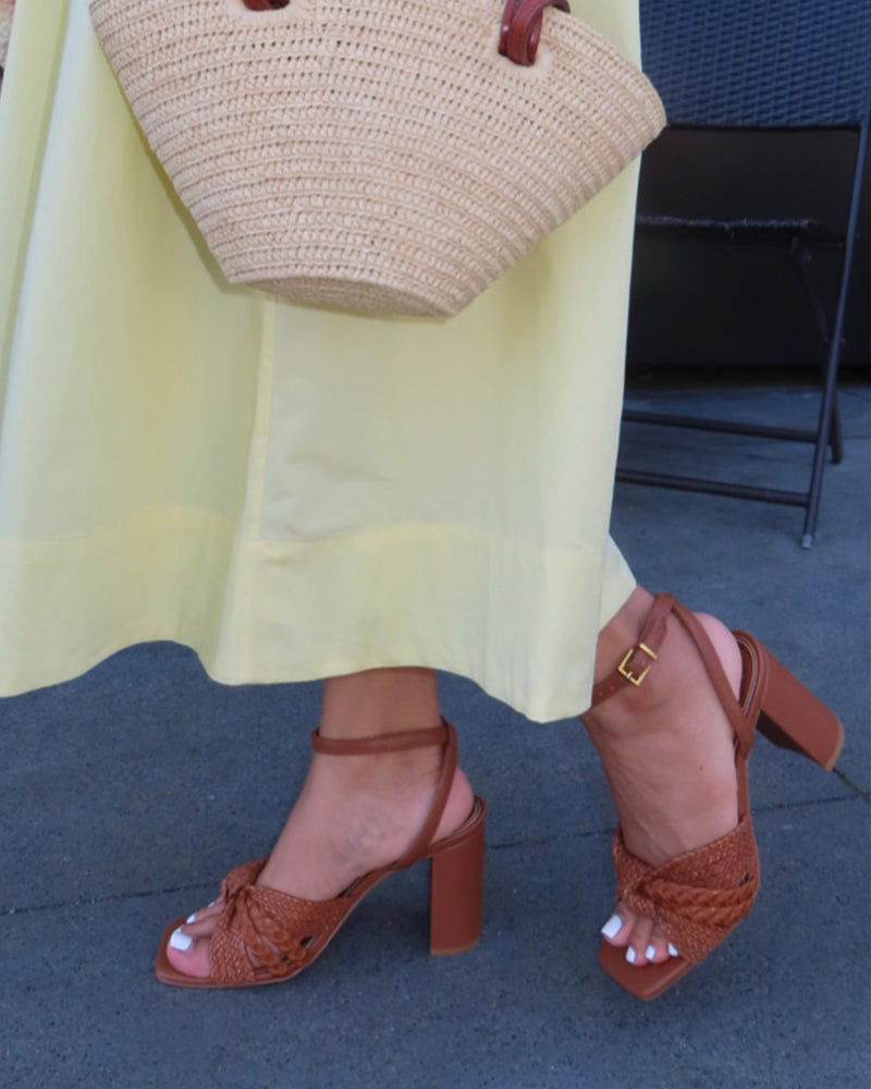 A person stands on gray pavement wearing a light yellow dress and Vince Camuto’s Hanlea Sandal, paired with a large straw handbag. Their toenails are painted white.