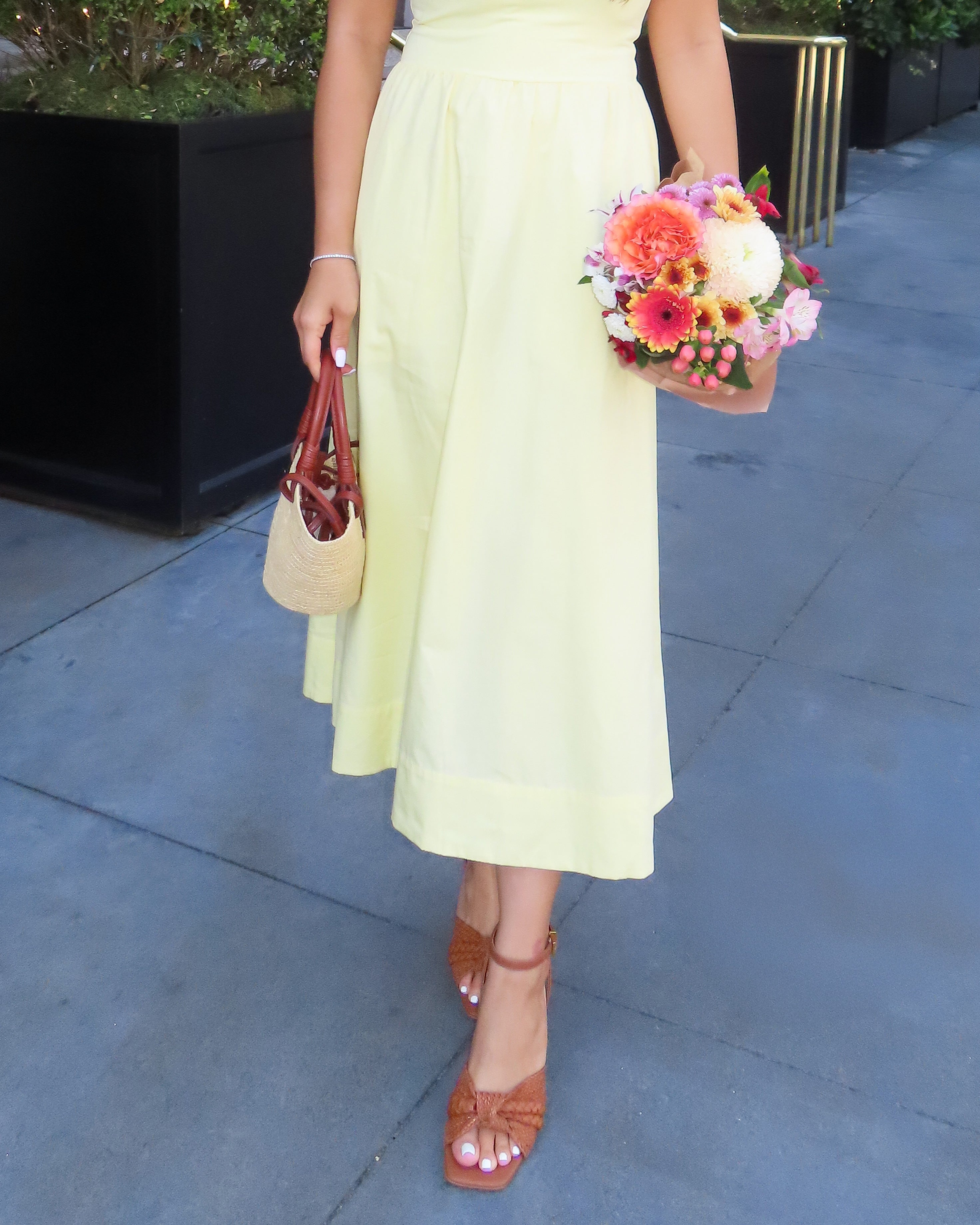 A woman in a pale yellow dress with a small bouquet and straw handbag wears Vince Camuto Hanlea Sandals, standing on a sidewalk.