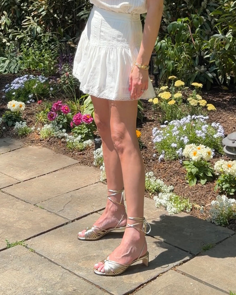 A person stands on a sunny stone patio near blooming flowers, wearing a white skirt, white top, and Vince Camuto Lory Kitten Heel Sandals with ankle ties. Their face is not visible.