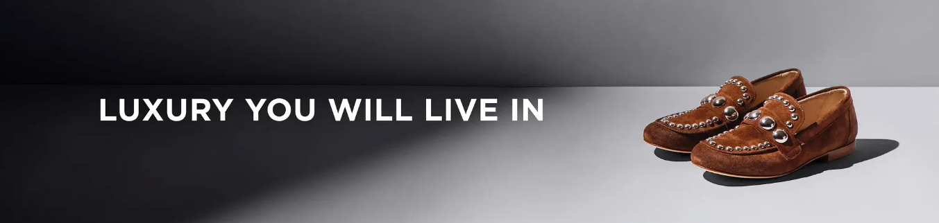 A pair of brown suede loafers with silver studs is displayed on a two-tone surface. Bold white text on the left reads, LUXURY YOU WILL LIVE IN.