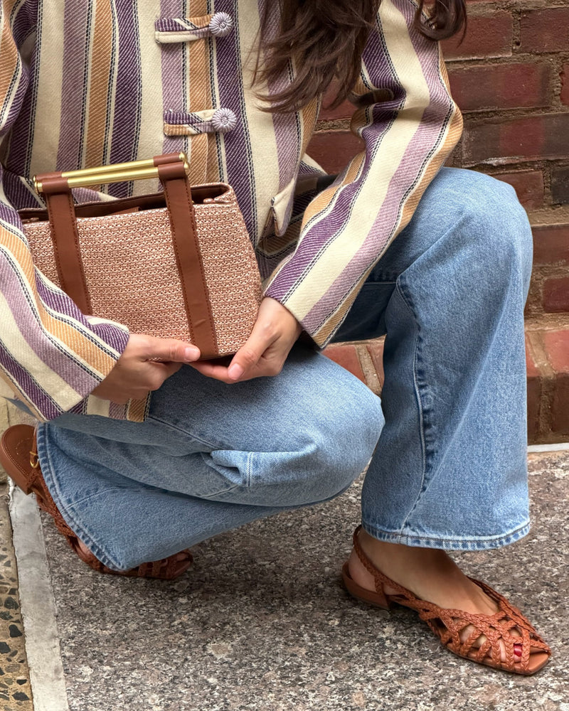 A person in light blue jeans, a striped blazer, and Vince Camuto's Stiorra Sandals kneels on the pavement, holding a textured beige and brown handbag near a brick wall.