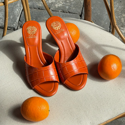 A pair of orange heeled sandals with a textured pattern sit on a beige cushioned chair, surrounded by three whole oranges.