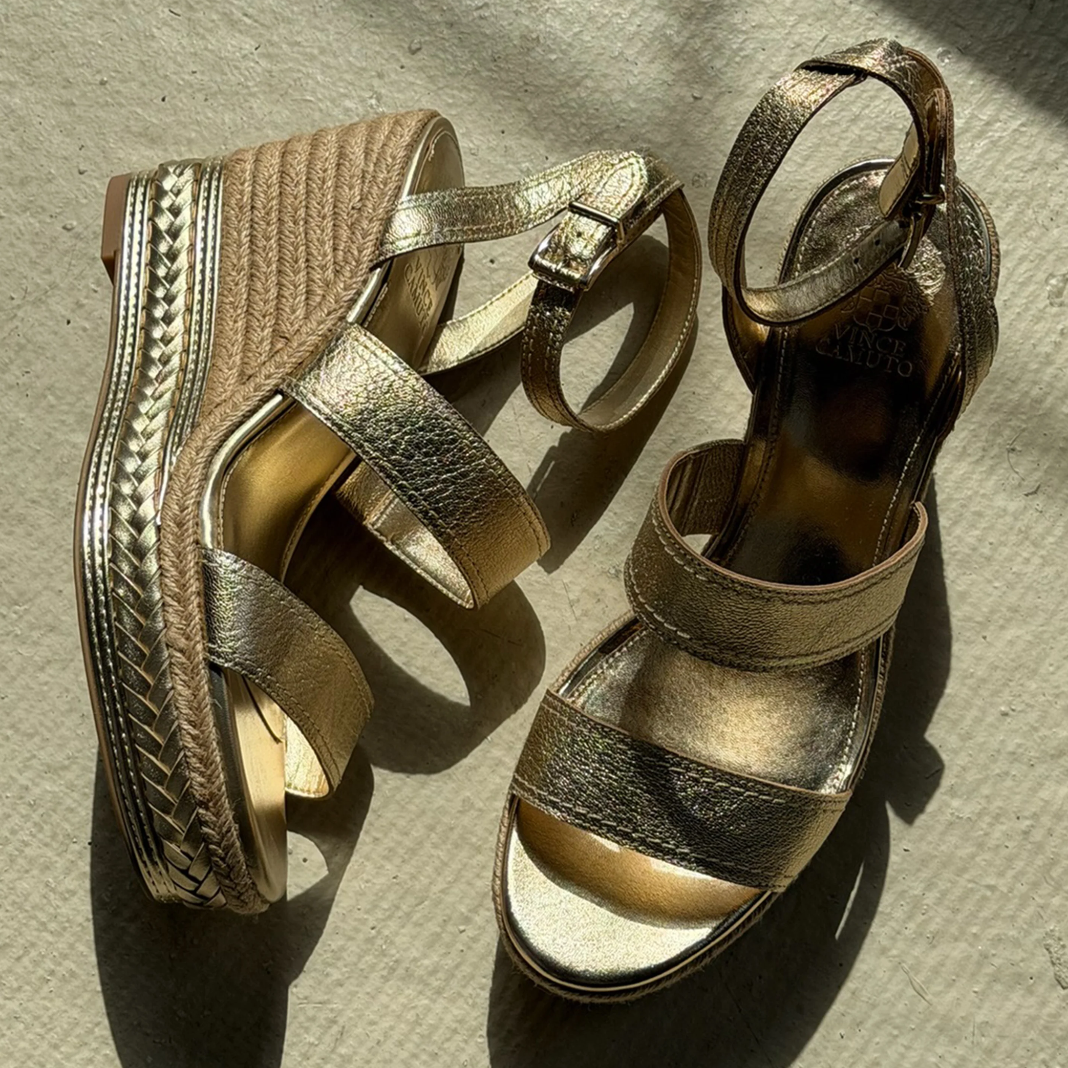 A pair of gold strappy wedge sandals with braided jute soles and metallic detailing, placed on a textured beige surface in sunlight.