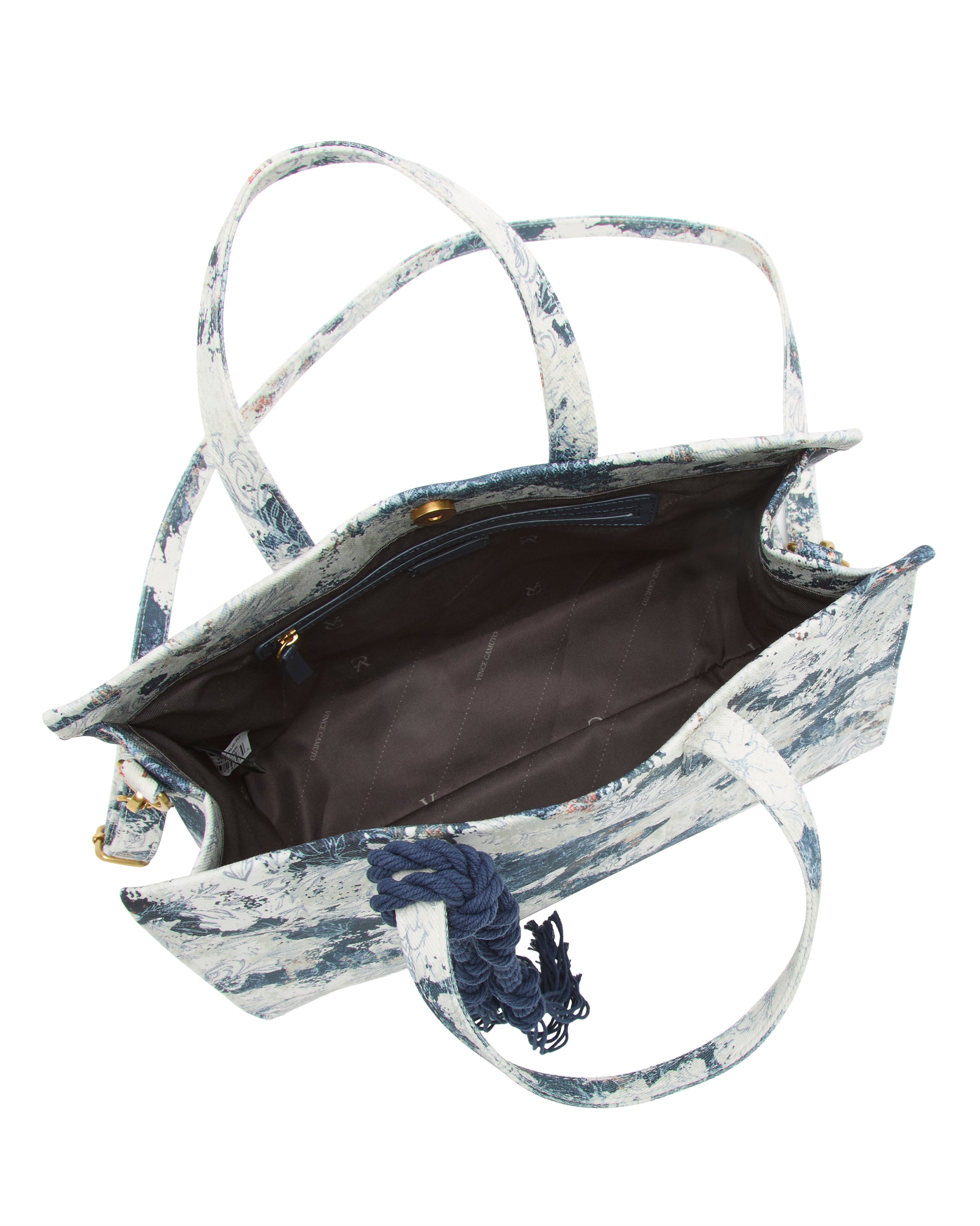 A top view of the Vince Camuto Aalis Tote Bag reveals a blue and white patterned canvas design with two handles, a blue tassel accent, and a roomy brown interior with a zippered pocket.