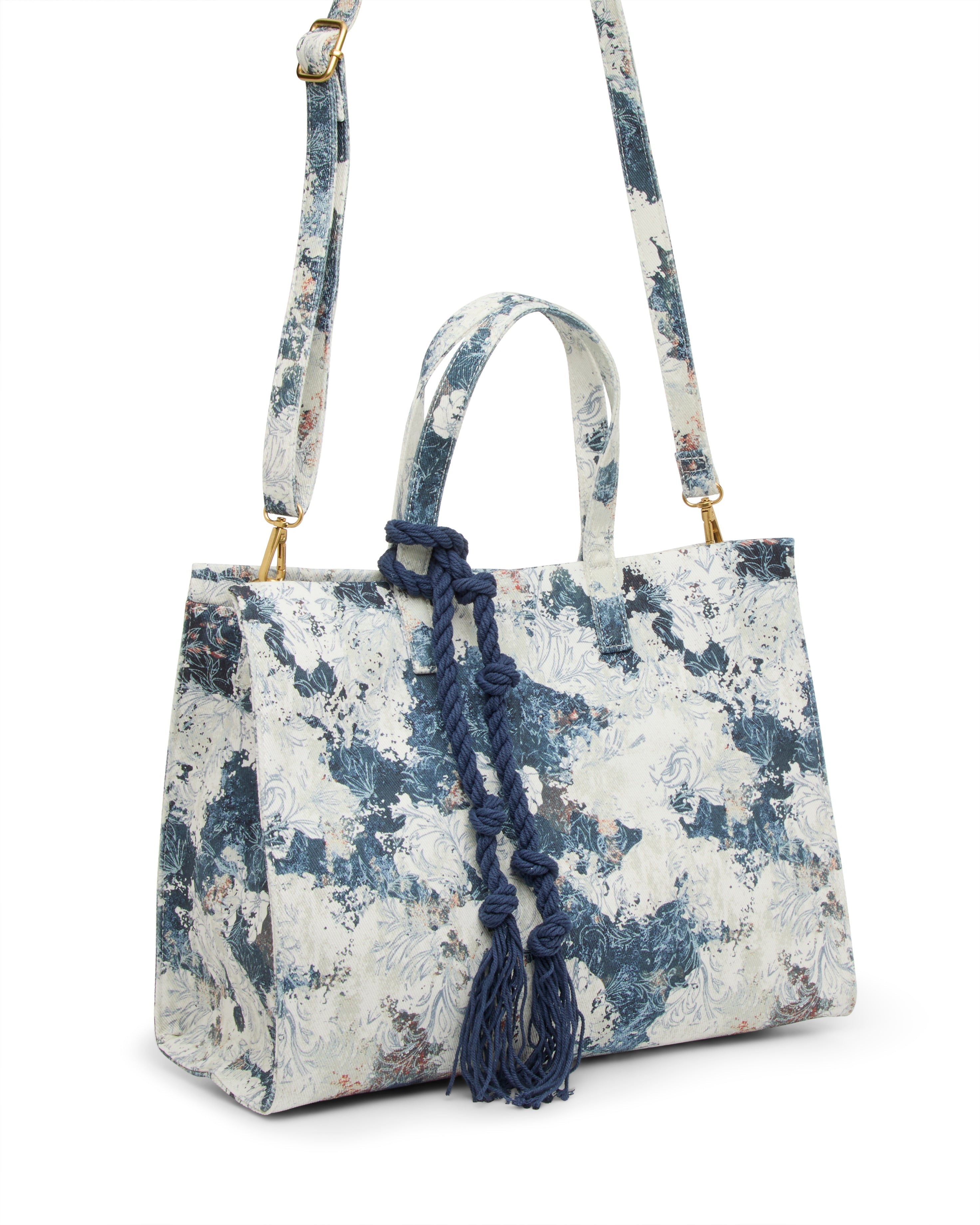 The Vince Camuto Aalis Tote Bag is a structured canvas handbag with a blue and white abstract floral pattern, top handle, adjustable shoulder strap, and a decorative navy knotted cord hanging from one handle.
