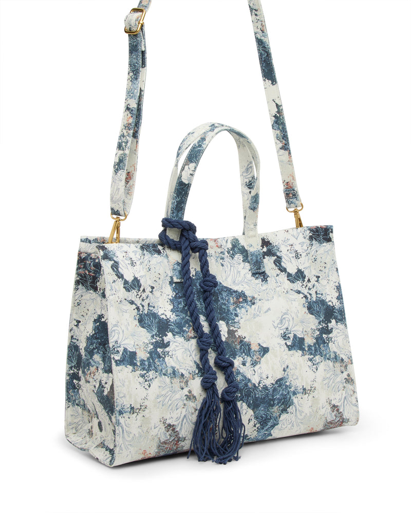The Vince Camuto Aalis Tote Bag is a structured canvas handbag with a blue and white abstract floral pattern, top handle, adjustable shoulder strap, and a decorative navy knotted cord hanging from one handle.