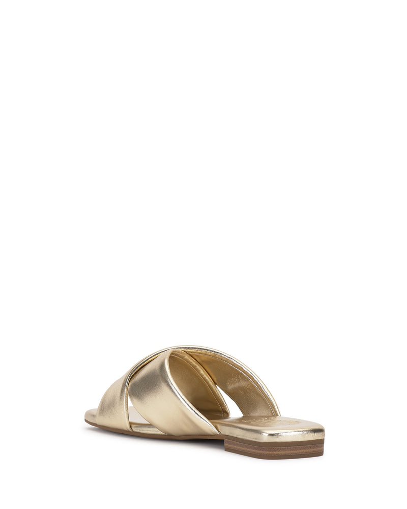 The Vince Camuto Adisey Cross-Band Sandal features gold metallic crisscross straps and a flat brown sole for everyday comfort. Shown from a back angle on a white background.