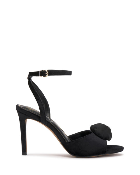 The Vince Camuto Aisven Sandal is a black high-heeled sandal featuring an ankle strap, open toe, and a decorative bow at the front. With its thin elegant heel and minimalist, sophisticated design, this shoe is the perfect blend of chic style and refinement.