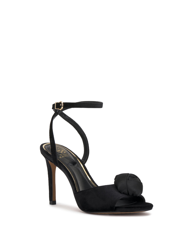 The Vince Camuto Aisven Sandal is a black high-heeled sandal featuring an ankle strap, open toe, and a decorative flower detail on the front strap. It has a slim heel and an inner sole adorned with gold Vince Camuto branding.