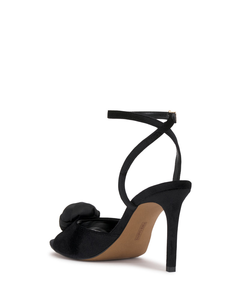 The Aisven Sandal by Vince Camuto is a black high-heeled sandal featuring an ankle strap and a bow detail on the front, presented from a rear angle against a white background.