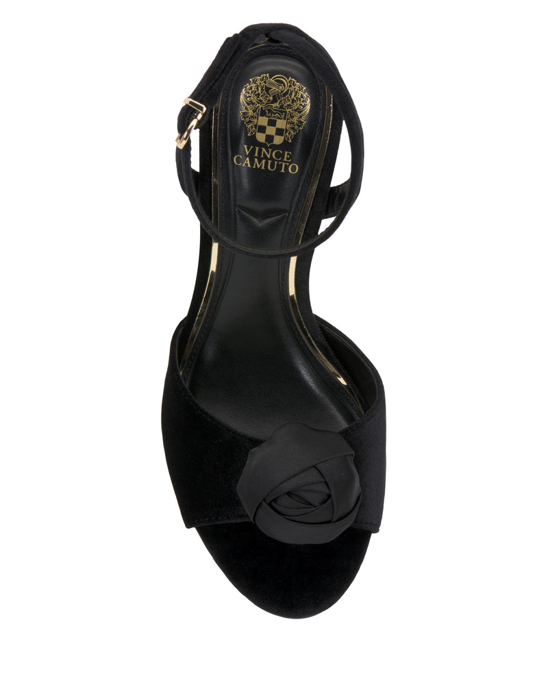 The Aisven Sandal by Vince Camuto is a black women’s sandal featuring an ankle strap, open toe, and a decorative fabric flower on the toe. The insole displays the gold Vince Camuto logo.