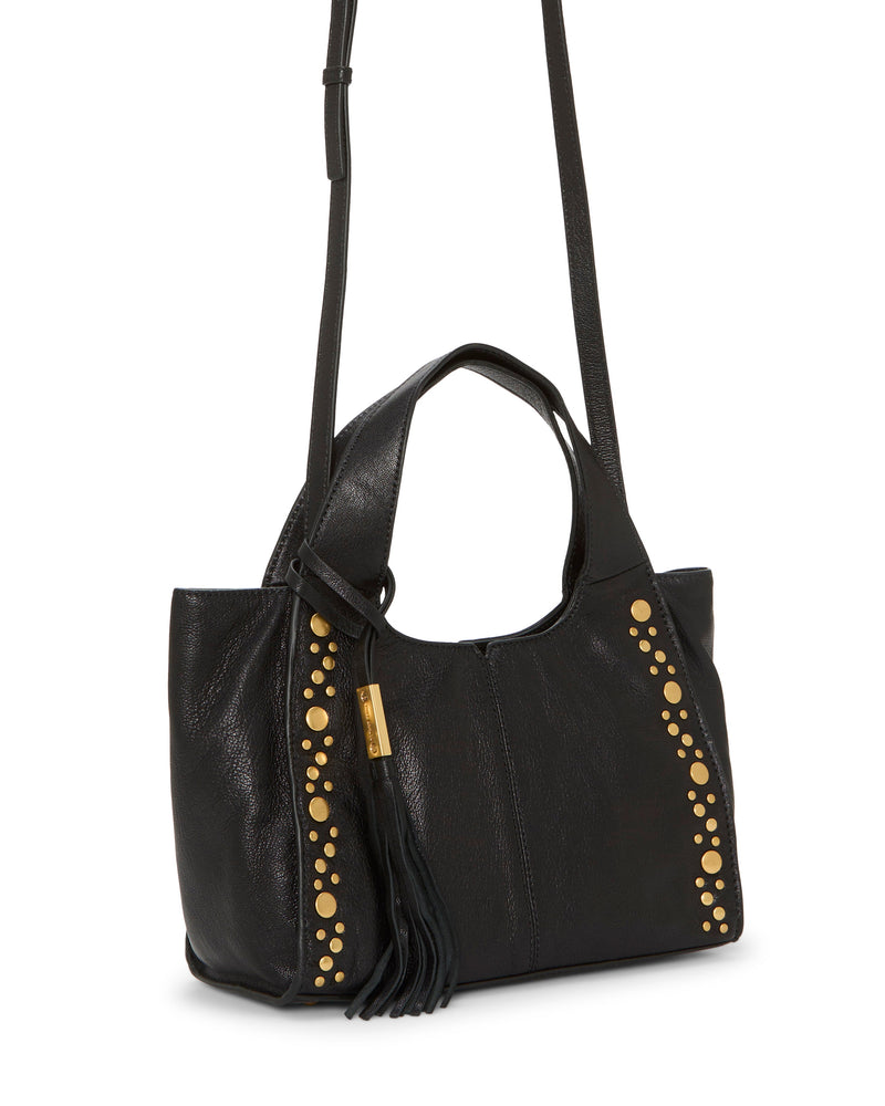 The Aleah Satchel Bag by Vince Camuto is crafted from black leather and features two handles, an adjustable long crossbody strap, gold stud accents on the front, and a black tassel keychain detail hanging at the side.