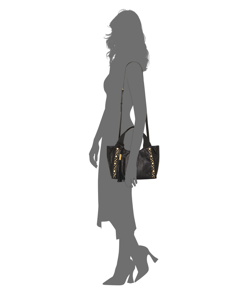 A woman in a dress and heels carries the Vince Camuto Aleah Satchel Bag, featuring gold stud accents, a tassel detail, and a versatile crossbody strap, over her shoulder against a white background.