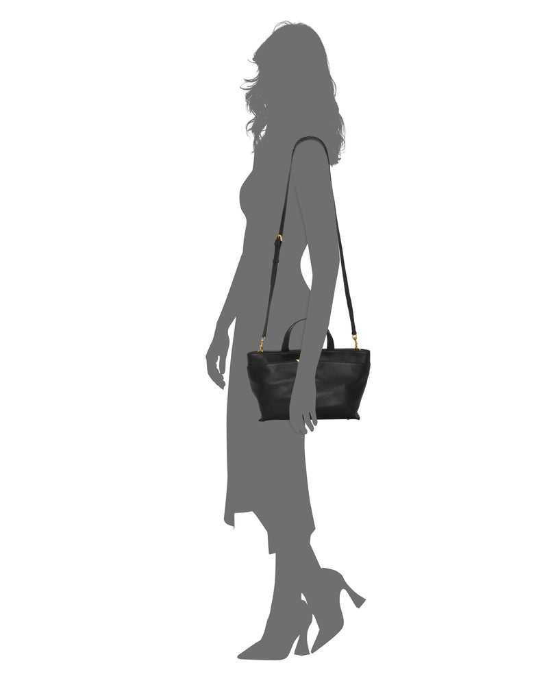 The Vince Camuto April Satchel Bag, featuring black leather and gold hardware, is shown hanging from the shoulder of a silhouetted woman in a long dress and heels, posed in profile against a white background.