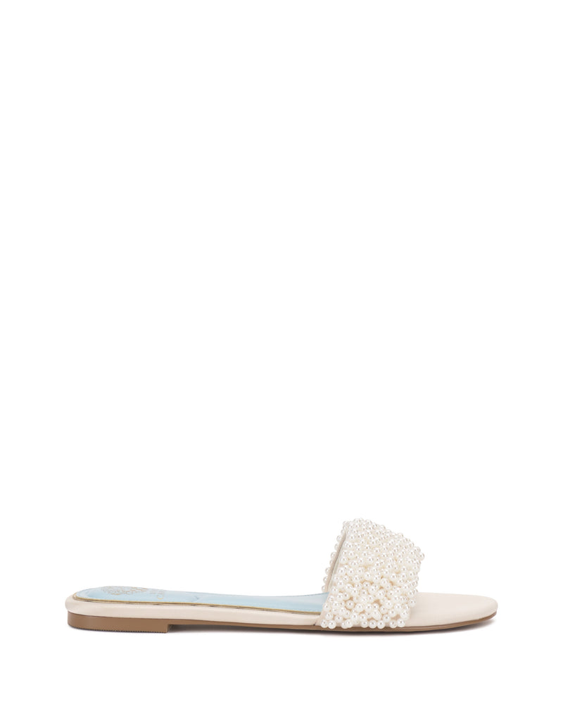 The Vince Camuto Balea Beaded Sandal features a beige sole and a wide strap adorned with elegant white pearl beading, creating a versatile and stylish flat sandal against a plain white background.