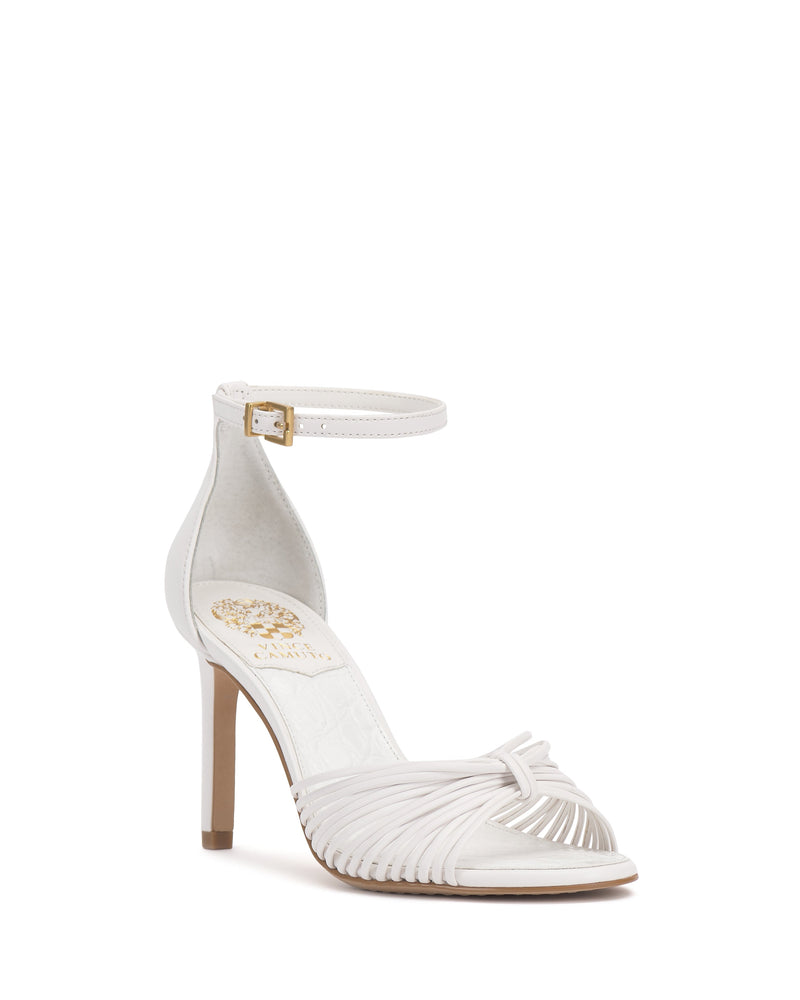The Blaine Sandal by Vince Camuto features a white strappy design, tall stiletto heel, open toe, ankle strap with gold buckle, and a gold emblem on the insole.