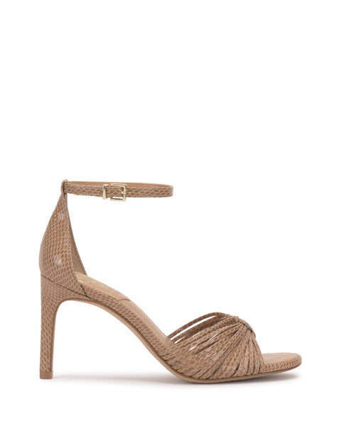 The Blaine Sandal by Vince Camuto features a tan, reptile-embossed design with twisted front straps, gold buckle ankle strap, open toe, and a sleek stiletto heel.
