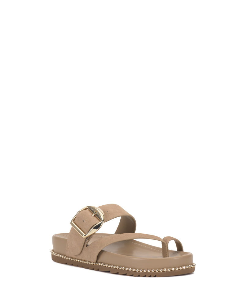 The Camilo Slide by Vince Camuto is a beige sandal with a wide buckle strap, large gold buckle, toe loop, and flat sole accented by small metallic studs along the edge.