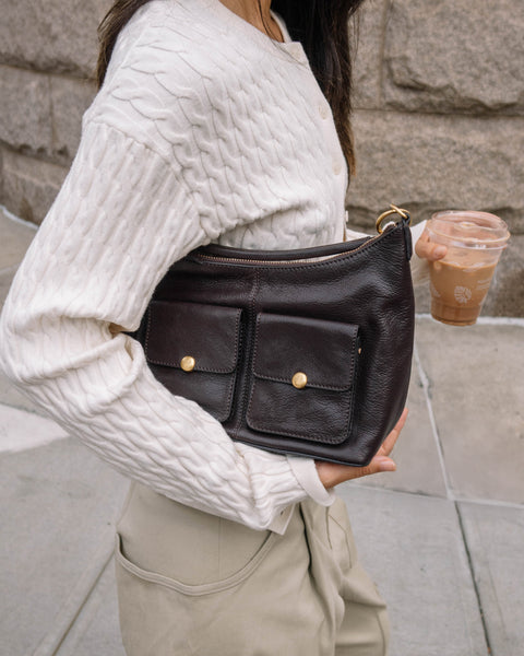 A woman in a white cable-knit sweater and beige pants carries the Vince Camuto Carly Shoulder Bag with two front pockets, gold buttons, and a crossbody strap as she sips iced coffee while walking outdoors.