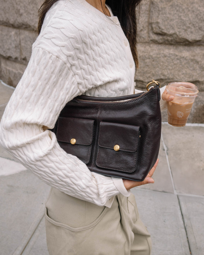 A woman in a white cable-knit sweater and beige pants carries the Vince Camuto Carly Shoulder Bag with two front pockets, gold buttons, and a crossbody strap as she sips iced coffee while walking outdoors.