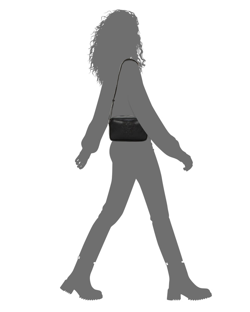 Silhouette of a person walking with the Vince Camuto Dalle Crossbody Bag, featuring a distinctive logo webbing strap slung over their shoulder, wearing boots and long pants, against a plain white background.
