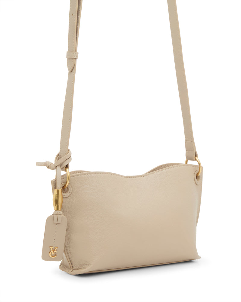 The Vince Camuto Ellah Crossbody Bag is a small beige leather bag with gold hardware, an adjustable strap, and a matching beige tag with a gold logo charm. Its simple, elegant design features a subtly wavy top edge.