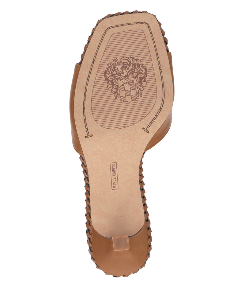 The image shows the sole of a tan Vince Camuto Emmla Mule with a square toe, braided sides, and a floral emblem with a shield center plus a rectangular HAND MADE mark, reflecting its modern elegance.
