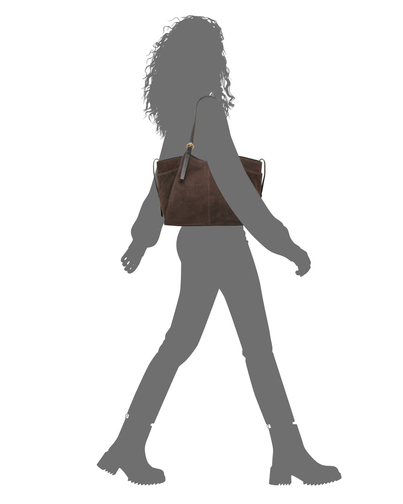 A silhouette of a person walking with the Vince Camuto Faey Tote Bag, a large dark brown purse featuring a keychain or tag on the strap, carried on their shoulder. The person is wearing boots.