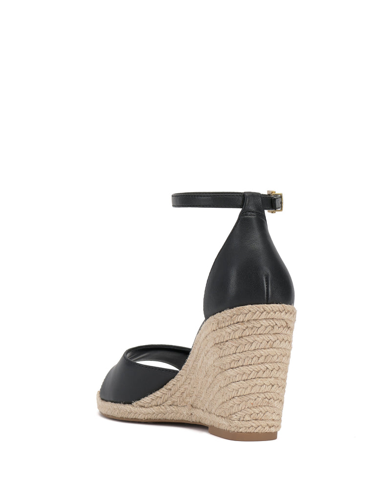 The Vince Camuto Felyn Espadrille Wedge Sandal is a black wedge with an ankle strap and buckle, featuring a high braided jute espadrille heel and platform, shown from the back against a white background.
