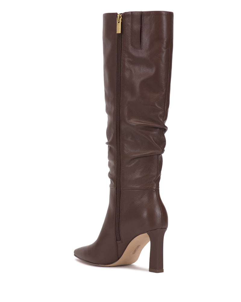 The Genisa Narrow Calf Slouchy Knee High Boot by Vince Camuto is a tall brown leather boot featuring a pointed toe, side zipper, and slightly slouchy shaft, displayed against a white background.