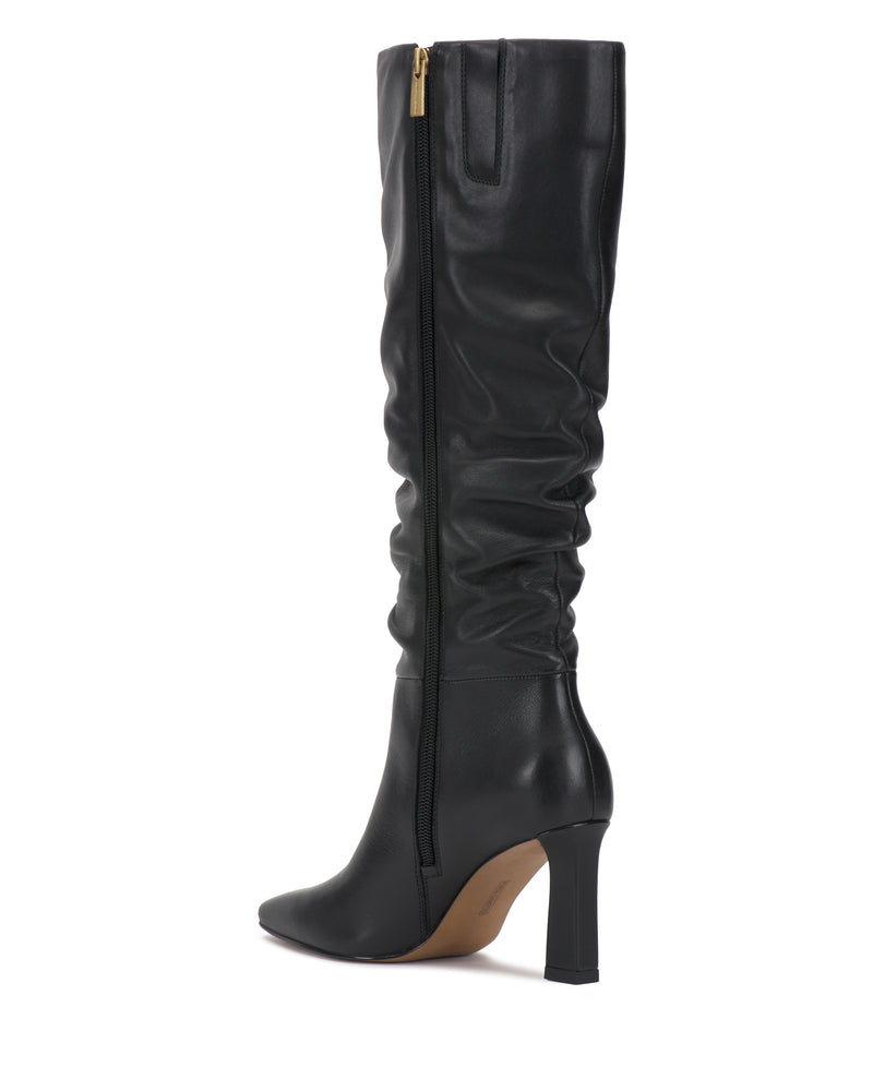 The Genisa Slouchy Knee High Boot by Vince Camuto features a black leather shaft with a slightly slouched silhouette, a pointed toe, and a mid-high block heel. Designed with a light brown sole and accented by a gold zipper at the top, this boot is pictured against a white background.