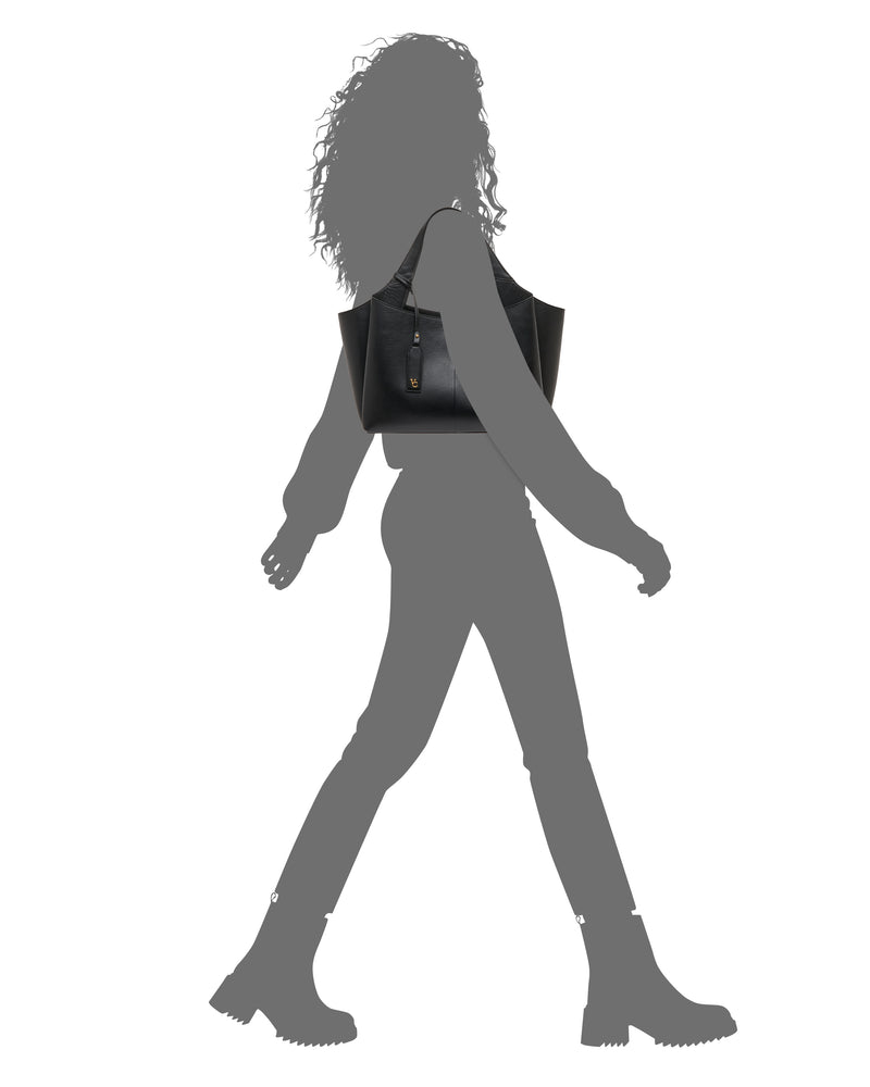 Silhouette of a person walking, wearing boots, with the Vince Camuto Gilda Tote Bag slung over their shoulder against a white background.