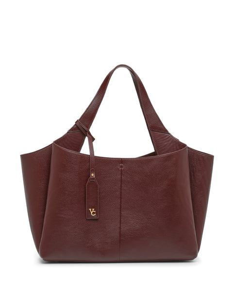 A large Vince Camuto Gilda Tote Bag in burgundy leather features double handles and a gold VG logo tag hanging from one strap, showcased against a white background.