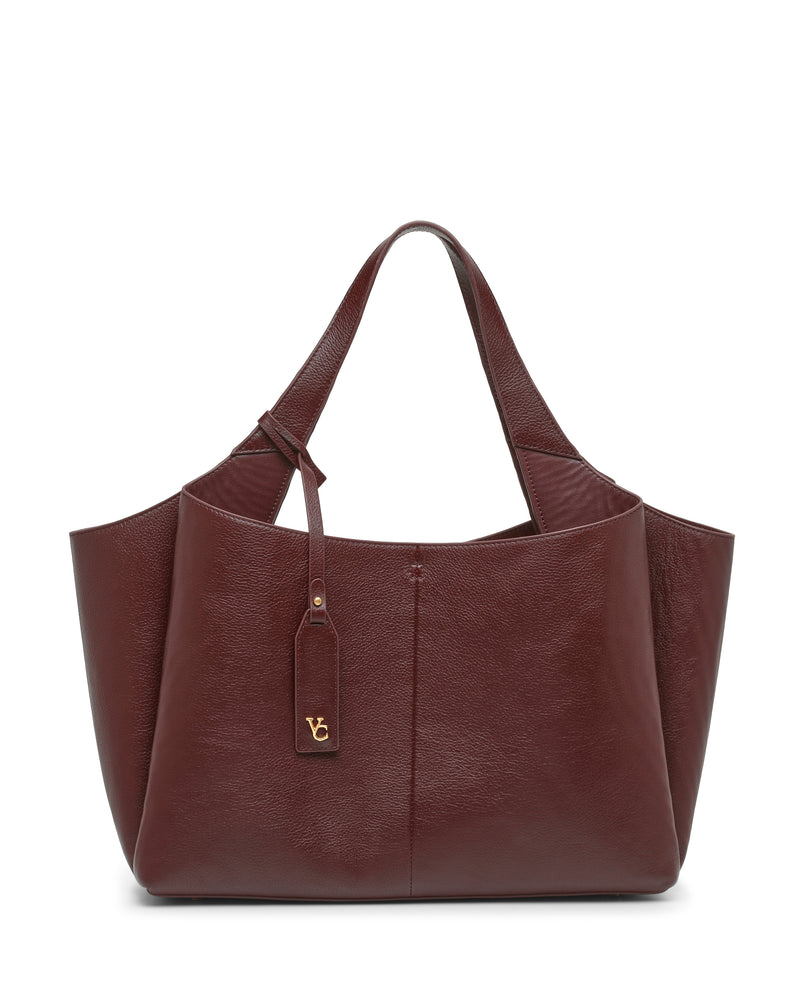 A large Vince Camuto Gilda Tote Bag in burgundy leather features double handles and a gold VG logo tag hanging from one strap, showcased against a white background.