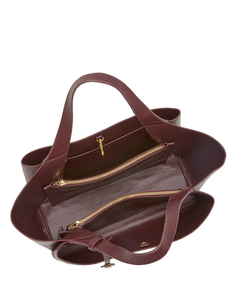 The Vince Camuto Gilda Tote Bag in burgundy leather is shown from above, highlighting its dual handles, two gold-zippered compartments, and an interior pocket. With its structured silhouette and smooth texture, this stylish handbag stands out as a sophisticated accessory.