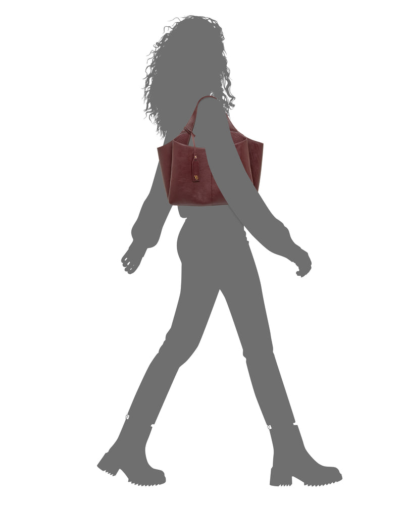 A silhouette of a person walking in boots carries the Vince Camuto Gilda Tote Bag—an elegant, sophisticated tote in rich brown leather—over their shoulder, set against a white background.