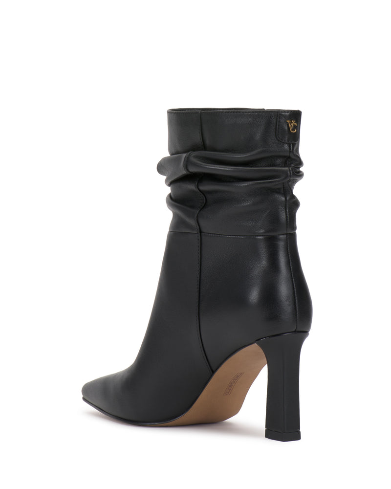 The Grenna Slouchy Bootie by Vince Camuto is a black leather ankle boot featuring a pointed toe, slouchy shaft, and a gold logo detail near the top, displayed on a white background.