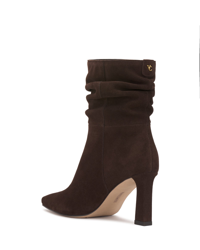 The Grenna Slouchy Bootie by Vince Camuto is a single brown suede ankle boot featuring a pointed toe, a slouchy upper, and a gold anchor emblem near the top of the shaft, shown from the side against a white background.