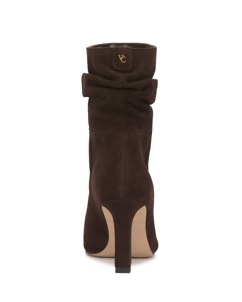 Back view of the Vince Camuto Grenna Slouchy Bootie in brown suede, showcasing a slightly slouched upper and a gold VC logo detail near the top. The boot features a beige sole and heel, set against a plain white background.