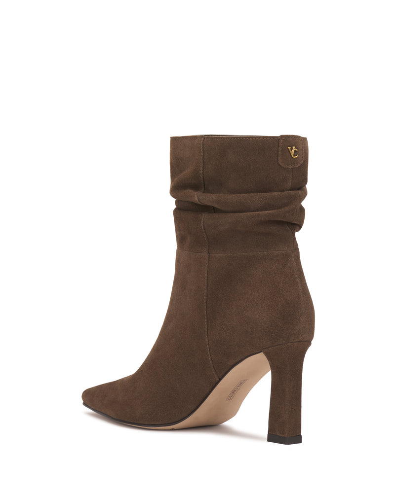 The Grenna Slouchy Bootie by Vince Camuto features brown suede with a slouchy design, a pointed toe, and a small gold Vince Camuto logo on the upper side. It has a slim high heel with a black tip, while the interior and sole are light beige.