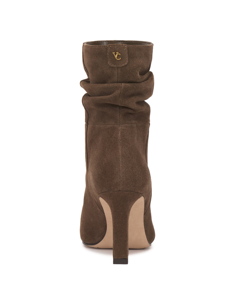 The Grenna Slouchy Bootie by Vince Camuto is a brown suede heeled boot viewed from the back, featuring a relaxed slouchy shaft and a small gold VC logo near the top. The light tan sole and heel are complemented by a center seam running down the back.